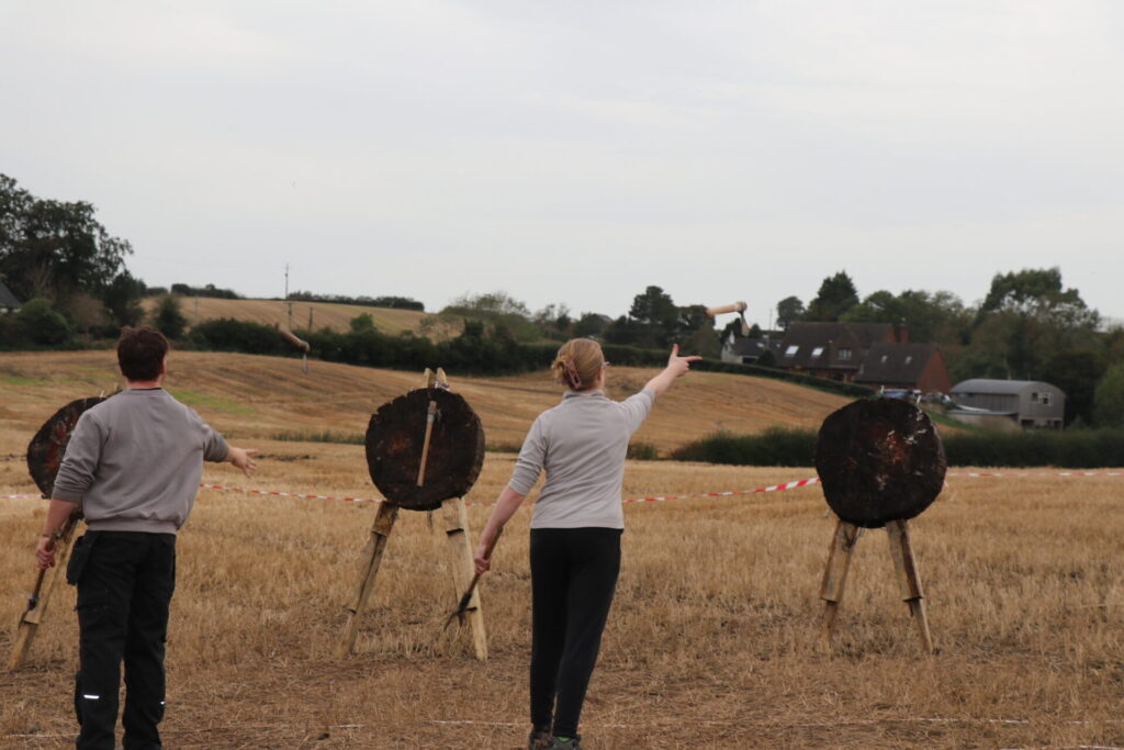 Two people (black trousers, grey tops) are throwing axes at wooden targets on a stubble field. In the background are hedgerows and a brown house.