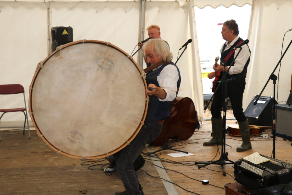 Willie Drennan (white shirt, black waistcoat, black trousers) playing a big drum hung on his shoulders. There are two people dressed the same playing the guitar in the background. They are inside a marquee with a wooden floor.