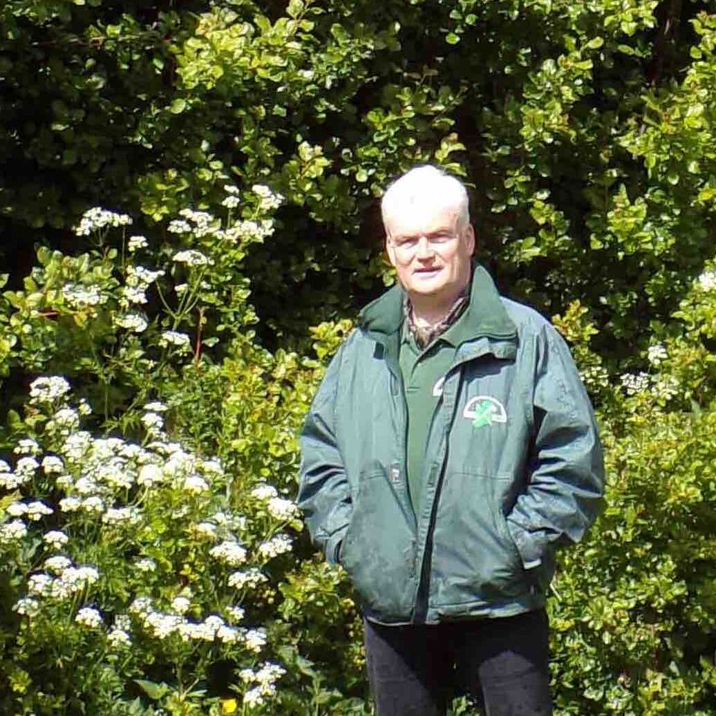 A photo of Mark McDowell standing in front of a hedgerow. He is a white man with white hair, wearing a green jumper and jacket with black trousers.