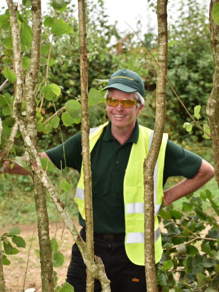 A photo of Adrian Fitzpatrick standing in between tree stems with a hedgerow in the background. He is a white man wearing a green cap, glasses, dark green t-shirt and a hi-vis. 