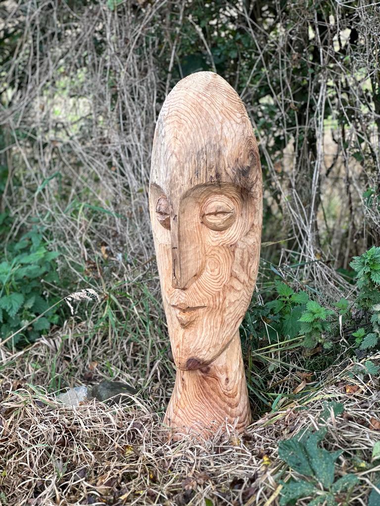A wooden sculpture of a face is on display in front of a hedgerow.