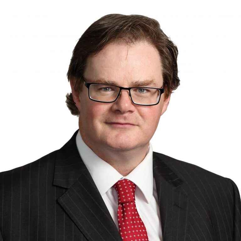 A headshot of Shane Downer. He is a white man with short brown hair. He is wearing glasses, a black suit, white shirt and red tie.
